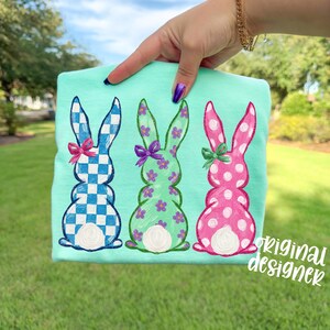 May include: A light teal t-shirt featuring three embroidered Easter bunnies. The bunnies are in blue and white checkered, green with floral accents, and pink with white polka dots. Each bunny has a bow. The text "original designer" is in the lower right corner.
