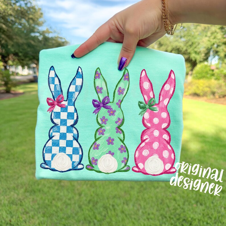 May include: A light teal t-shirt featuring three embroidered Easter bunnies. The bunnies are in blue and white checkered, green with floral accents, and pink with white polka dots. Each bunny has a bow. The text "original designer" is in the lower right corner.
