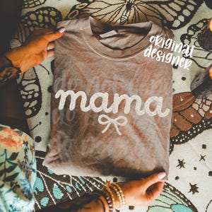 May include: A brown t-shirt with the word "mama" in white, rope-like lettering and a small bow. The words "do not" are faintly visible above and below "mama". The shirt is folded and held by hands.