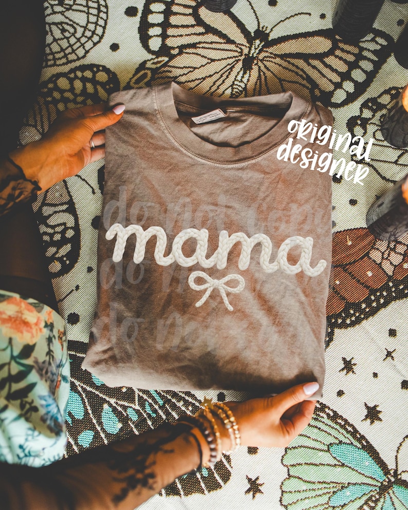 May include: A brown t-shirt with the word "mama" in white, rope-like lettering and a small bow. The words "do not" are faintly visible above and below "mama". The shirt is folded and held by hands.