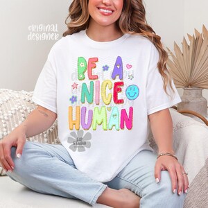 May include: White t-shirt with the phrase "BE A NICE HUMAN" in colorful bubble letters. The design includes flowers, stars, and a smiley face. The shirt is a casual, comfortable style.