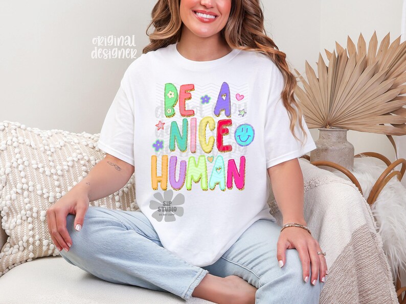 May include: White t-shirt with the phrase "BE A NICE HUMAN" in colorful bubble letters. The design includes flowers, stars, and a smiley face. The shirt is a casual, comfortable style.