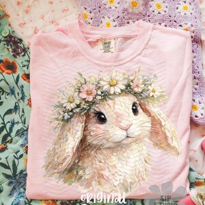 May include: A light pink t-shirt featuring a detailed illustration of a bunny wearing a floral crown. The bunny has large eyes and soft fur. The text "original designer" is printed at the bottom of the shirt.