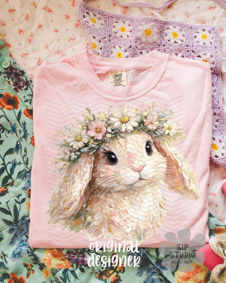 May include: A light pink t-shirt featuring a detailed illustration of a bunny wearing a floral crown. The bunny has large eyes and soft fur. The text "original designer" is printed at the bottom of the shirt.