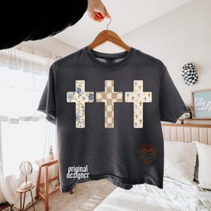 May include: A charcoal gray short-sleeve t-shirt featuring a graphic of three crosses. The left cross has a blue floral pattern, the center cross has a beige checkered pattern, and the right cross has a blue and beige pattern with small crosses.
