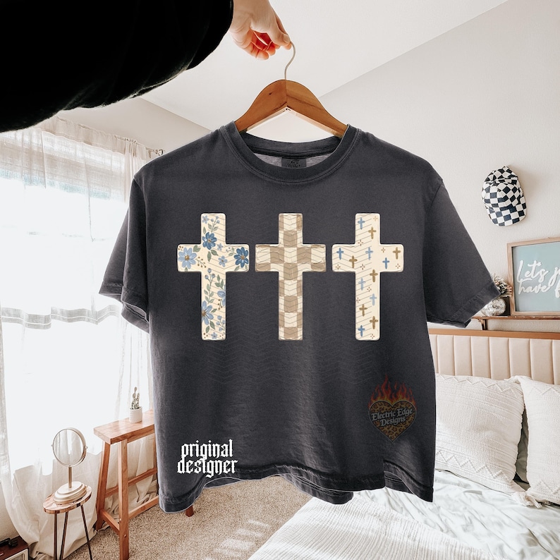 May include: A charcoal gray short-sleeve t-shirt featuring a graphic of three crosses. The left cross has a blue floral pattern, the center cross has a beige checkered pattern, and the right cross has a blue and beige pattern with small crosses.