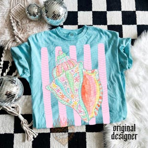 May include: A light blue t-shirt featuring a large, colorful seashell design with pink vertical stripes. The seashell is detailed with various sea creatures and patterns. The words "original designer" are printed on the bottom right.
