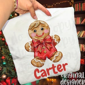 May include: White pillow with a gingerbread man design. The gingerbread man has a red bow tie and red outfit. The name "Carter" is printed in red script at the bottom. The words "Original Designer" are printed in green.