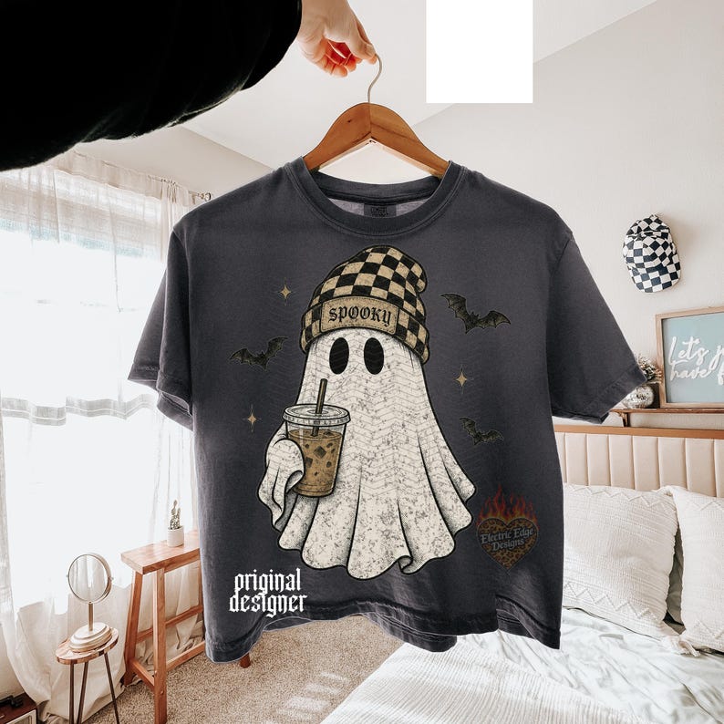 May include: A charcoal gray t-shirt featuring a graphic of a ghost wearing a checkered beanie and holding an iced coffee. The ghost is surrounded by small bats and stars. The text "SPOOKY" is on the beanie, and "original designer" is printed below.