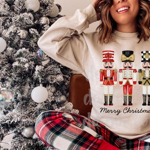 Peut inclure: Un sweat-shirt beige avec trois casse-noisettes illustr&eacute;s en uniformes rouges, noirs et dor&eacute;s. Les mots "Merry Christmas" sont &eacute;crits en dessous. Un sapin de No&euml;l avec des d&eacute;corations est en arri&egrave;re-plan.