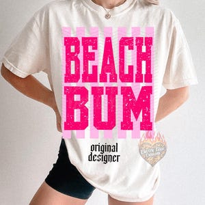 May include: Cream-colored t-shirt with the words "BEACH BUM" in distressed pink block letters. The shirt features pink striped accents and the text "original designer" in a script font. A heart-shaped logo is visible on the lower right.