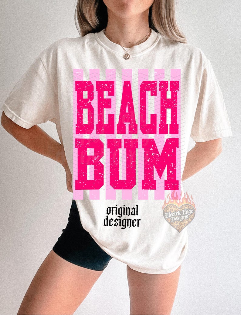 May include: Cream-colored t-shirt with the words "BEACH BUM" in distressed pink block letters. The shirt features pink striped accents and the text "original designer" in a script font. A heart-shaped logo is visible on the lower right.