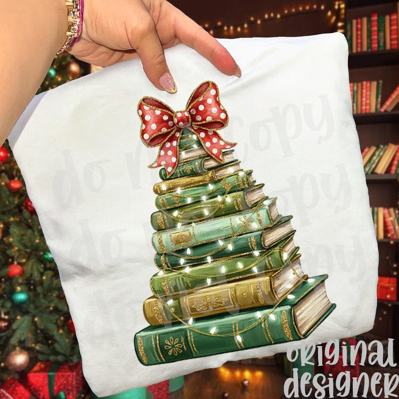 May include: White pillow with a Christmas tree design made of stacked books. The books are green and gold, adorned with string lights and topped with a red bow with white polka dots. The pillow is held by a hand.