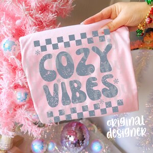 May include: A light pink t-shirt with the words "COZY VIBES" in a distressed gray font. The design includes gray checkerboard patterns and snowflakes. The words "original designer" are printed in a script font.