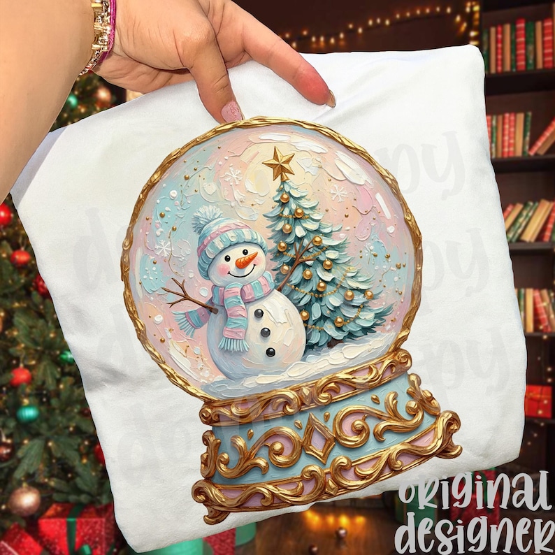 May include: A white t-shirt featuring a graphic of a snow globe with a snowman wearing a blue and pink striped hat and scarf, standing next to a decorated Christmas tree with a gold star topper, all set in an ornate gold and blue base.