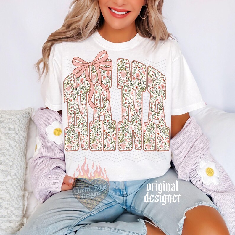 May include: White t-shirt with the word "NANA" in large, floral-patterned letters. A pink bow is above the word. The shirt has a relaxed fit and is suitable for casual wear. The design includes the text "Electric Edge Designs" and "original designer".
