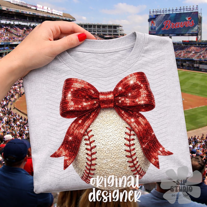 May include: A light gray t-shirt featuring a baseball with a red sequined bow. The baseball is white with red stitching. The text "Original Designer" is at the bottom of the shirt.