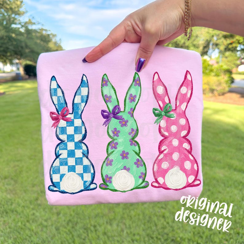 May include: A light pink t-shirt featuring three Easter bunny designs. The bunnies are in blue and white checkered, green with purple flowers, and pink with white polka dots. Each bunny has a bow. The text "original designer" is in the bottom right corner.