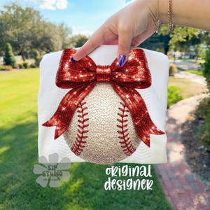 May include: White t-shirt featuring a baseball design with a red sequined bow. The baseball is covered in sequins and has red stitching. The text "original designer" is visible on the shirt.