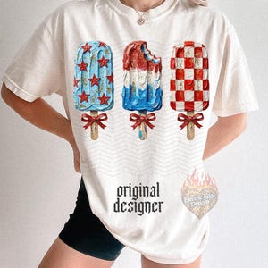 Pu&ograve; includere: T-shirt color crema con tre disegni di ghiaccioli rossi, bianchi e blu. Un ghiacciolo ha stelle rosse su sfondo blu, un altro &egrave; parzialmente mangiato e il terzo ha un motivo a scacchiera rosso e bianco. Il testo "original designer" &egrave; stampato.