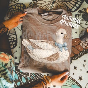 May include: A brown t-shirt featuring a patchwork duck design with a blue bow tie. The text "do not" is visible on the shirt. The words "original designer" are also visible on the shirt.