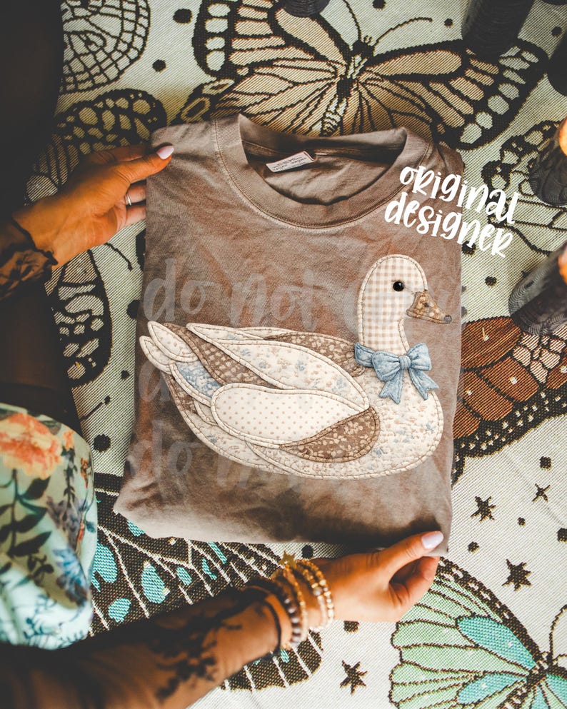 May include: A brown t-shirt featuring a patchwork duck design with a blue bow tie. The text "do not" is visible on the shirt. The words "original designer" are also visible on the shirt.