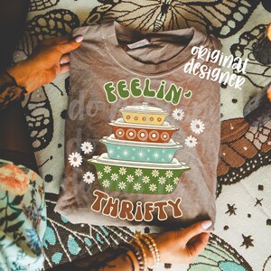 Pu&ograve; includere: Una maglietta marrone con le parole "FEELIN' THRIFTY" in verde e marrone. La maglietta presenta una grafica di pirofile colorate impilate e accenti di margherite bianche. La maglietta &egrave; esposta su una superficie a fantasia.
