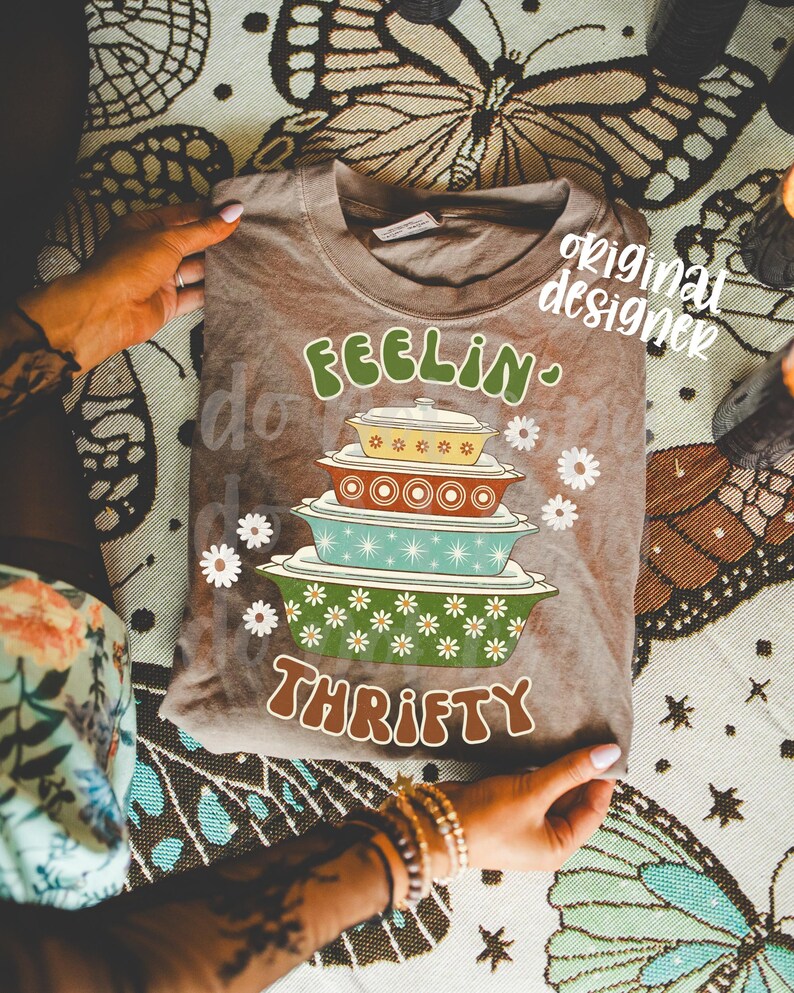 Pu&ograve; includere: Una maglietta marrone con le parole "FEELIN' THRIFTY" in verde e marrone. La maglietta presenta una grafica di pirofile colorate impilate e accenti di margherite bianche. La maglietta &egrave; esposta su una superficie a fantasia.