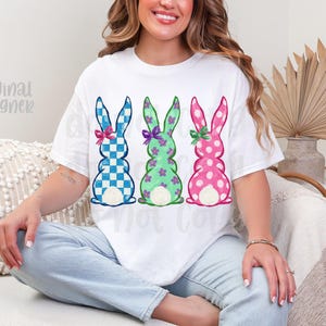May include: White t-shirt featuring three Easter bunny designs. The bunnies are in blue checkered, green floral, and pink polka dot patterns, each with a bow. The words "original designer" are visible on the left side of the shirt.