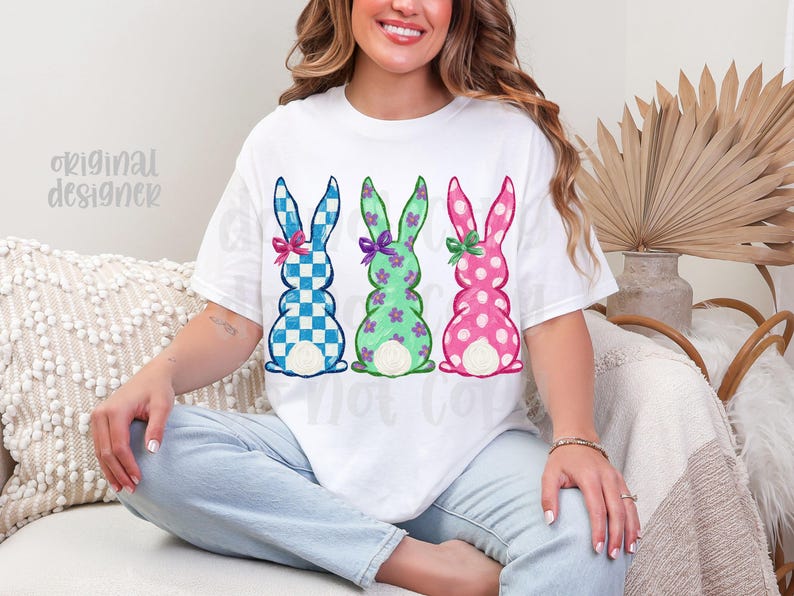 May include: White t-shirt featuring three Easter bunny designs. The bunnies are in blue checkered, green floral, and pink polka dot patterns, each with a bow. The words "original designer" are visible on the left side of the shirt.