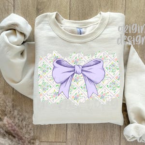 May include: A light beige sweatshirt featuring a large lavender bow design. The bow is centered on a floral and geometric pattern. The words "original designer" are printed on the upper right side of the sweatshirt.