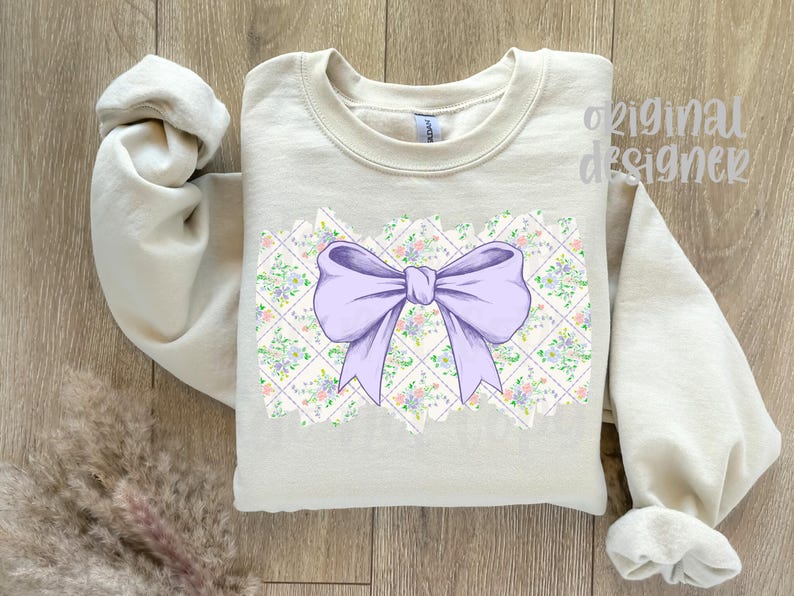 May include: A light beige sweatshirt featuring a large lavender bow design. The bow is centered on a floral and geometric pattern. The words "original designer" are printed on the upper right side of the sweatshirt.