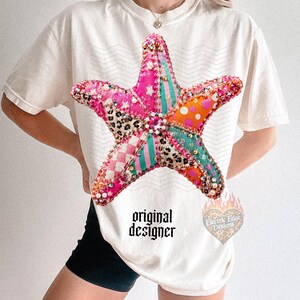 May include: Cream-colored t-shirt featuring a large, colorful starfish design. The starfish is made of various patterned fabrics and embellished with beads. The text "original designer" is printed below the starfish.