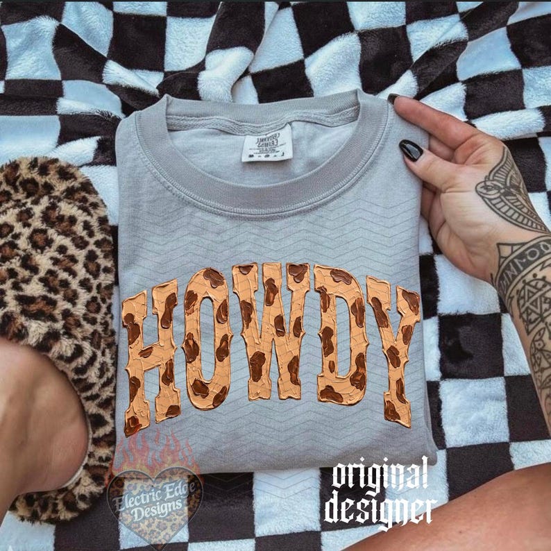 May include: A gray sweatshirt with the word "HOWDY" in a brown, textured font. The sweatshirt is on a black and white checkered blanket. A leopard print slipper is visible on the left.