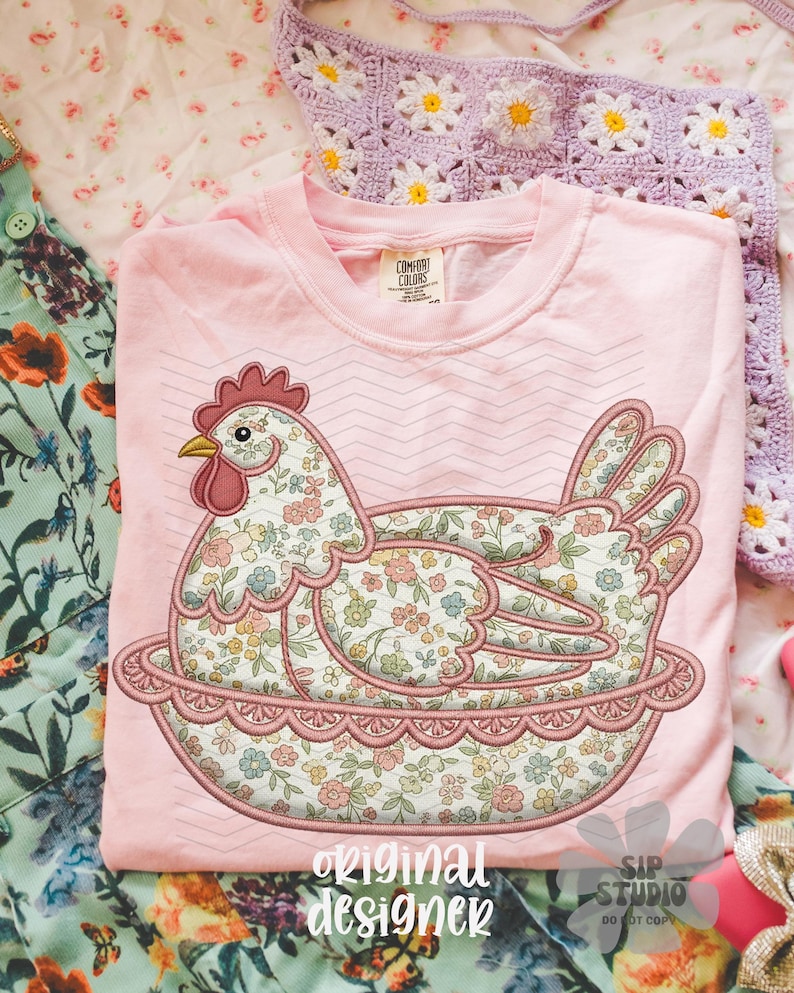 May include: A light pink t-shirt featuring a floral hen design. The hen is embroidered with a colorful floral pattern and sits in a decorative nest. The text "original designer" is printed at the bottom.