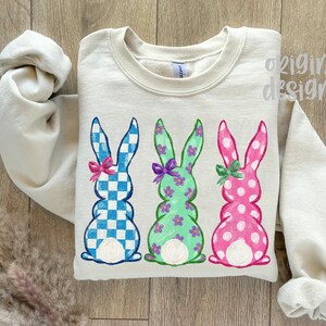 May include: A cream-colored sweatshirt featuring three Easter bunny designs. The bunnies are in blue and white checkered, green with purple flowers, and pink with white polka dots. Each bunny has a bow.