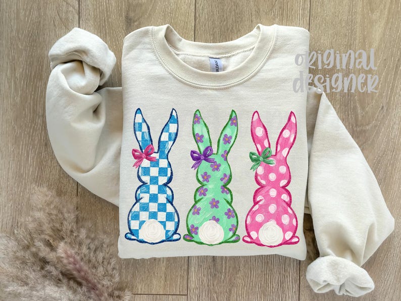 May include: A cream-colored sweatshirt featuring three Easter bunny designs. The bunnies are in blue and white checkered, green with purple flowers, and pink with white polka dots. Each bunny has a bow.