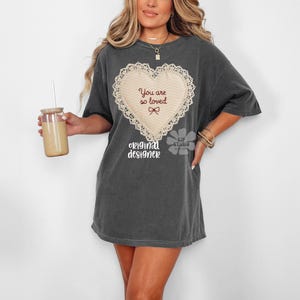 May include: Dark grey t-shirt dress with a cream heart design, the words "You are so loved" and a bow. The shirt also has the words "original designer" printed below the heart. A glass with a straw is held in the left hand.
