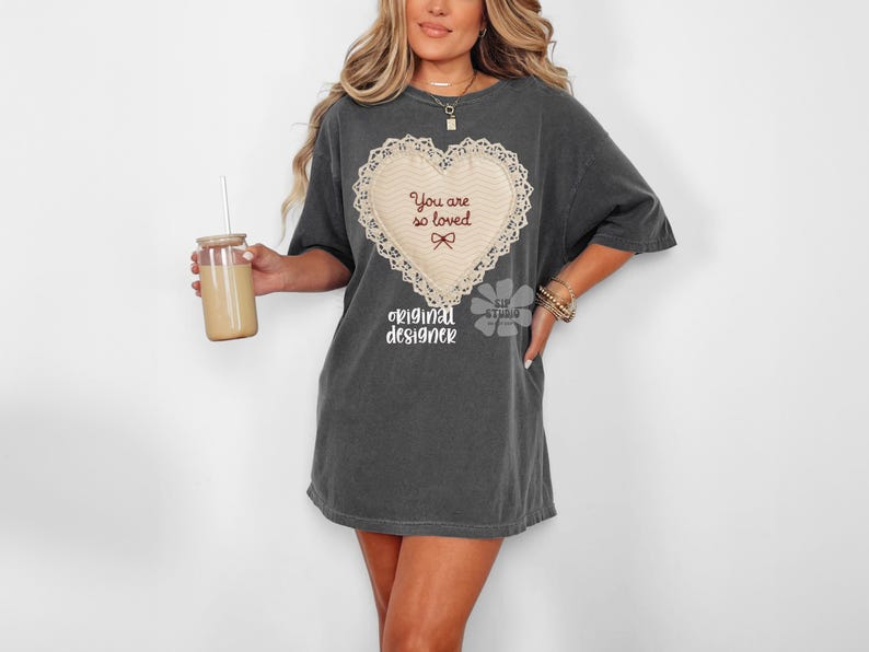 May include: Dark grey t-shirt dress with a cream heart design, the words "You are so loved" and a bow. The shirt also has the words "original designer" printed below the heart. A glass with a straw is held in the left hand.