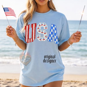 May include: Light blue t-shirt with the word "USA" in red and white striped, cherry, and blue checkered designs. The shirt has the text "original designer" and a logo. The person is holding a small American flag and a sparkler.
