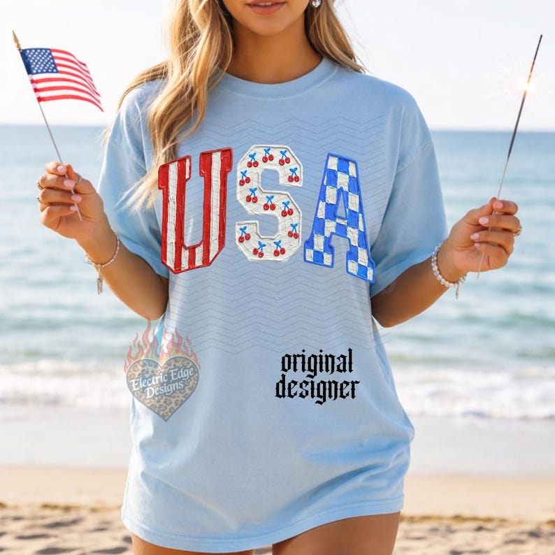 May include: Light blue t-shirt with the word "USA" in red and white striped, cherry, and blue checkered designs. The shirt has the text "original designer" and a logo. The person is holding a small American flag and a sparkler.