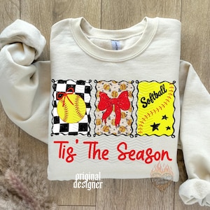 May include: A cream-coloured crewneck sweatshirt featuring a graphic with three rectangular panels. The panels display a yellow softball on a black and white checkerboard, a red bow over small softballs, and the word Softball with stars. The text Tis' The Season is printed in red below the panels.