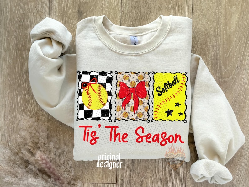 May include: A cream-coloured crewneck sweatshirt featuring a graphic with three rectangular panels. The panels display a yellow softball on a black and white checkerboard, a red bow over small softballs, and the word Softball with stars. The text Tis' The Season is printed in red below the panels.