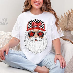 May include: White t-shirt featuring a Santa Claus graphic. Santa wears a red and black checkered hat with the word "Sleigh" and star-shaped sunglasses. The design has a festive, holiday theme.