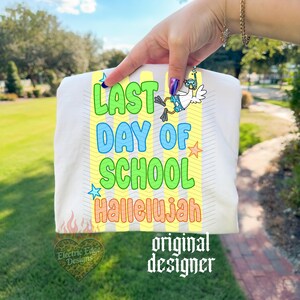 Puede incluir: Camiseta blanca con el texto "LAST DAY OF SCHOOL HALLELUJAH" en verde, azul y naranja. Tambi&eacute;n presenta un dibujo de un ganso con esn&oacute;rquel y aletas. La camiseta tiene un fondo a rayas amarillas y blancas.
