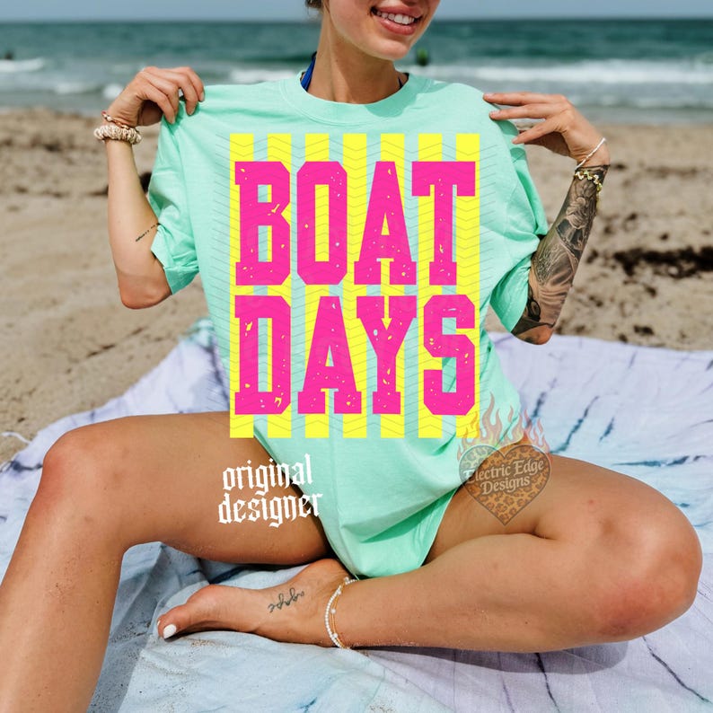 Może przedstawiać: Jasnozielona koszulka z napisem "BOAT DAYS" dużymi r&oacute;żowymi literami na ż&oacute;łtych paskach. Koszulka ma kr&oacute;tkie rękawy i jest noszona na plaży. Na koszulce wydrukowano słowa "original designer".