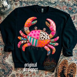 May include: Black sweatshirt featuring a colorful crab design with patchwork patterns. The crab has pink, orange, and green sections with star, leopard, and striped patterns. The text "original designer" is at the bottom.