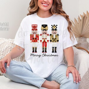 Peut inclure: T-shirt blanc avec trois illustrations de casse-noisettes festifs en rouge, blanc et or. Les mots "Merry Christmas" sont imprim&eacute;s en dessous. Le t-shirt est de style d&eacute;contract&eacute;.