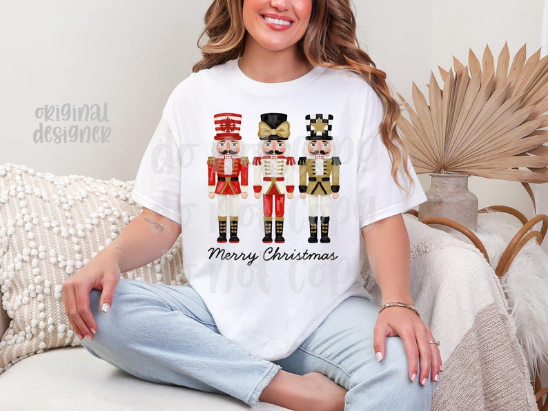Peut inclure: T-shirt blanc avec trois illustrations de casse-noisettes festifs en rouge, blanc et or. Les mots "Merry Christmas" sont imprim&eacute;s en dessous. Le t-shirt est de style d&eacute;contract&eacute;.