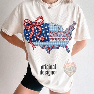 Pu&ograve; includere: T-shirt color crema con una grafica della mappa degli Stati Uniti con motivi patriottici. Il design include una sezione a quadretti blu e bianchi, una sezione con stelle e una sezione con cuori. Un fiocco rosso e bianco &egrave; sulla mappa. Le parole "original designer" sono stampate sulla maglietta.